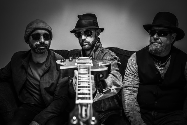 Three men wearing sunglasses hold a guitar in forced perspective.