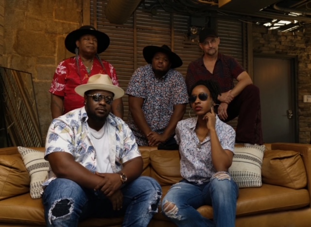 A group of five band members, two sitting on a brown sofa, three standing behind.